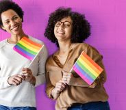 This is an image of two queer women holding the Pride flag on a pink wall.