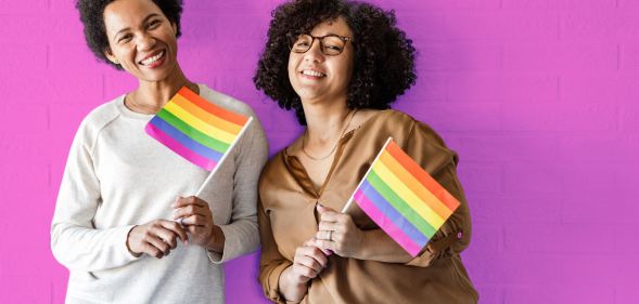 This is an image of two queer women holding the Pride flag on a pink wall.