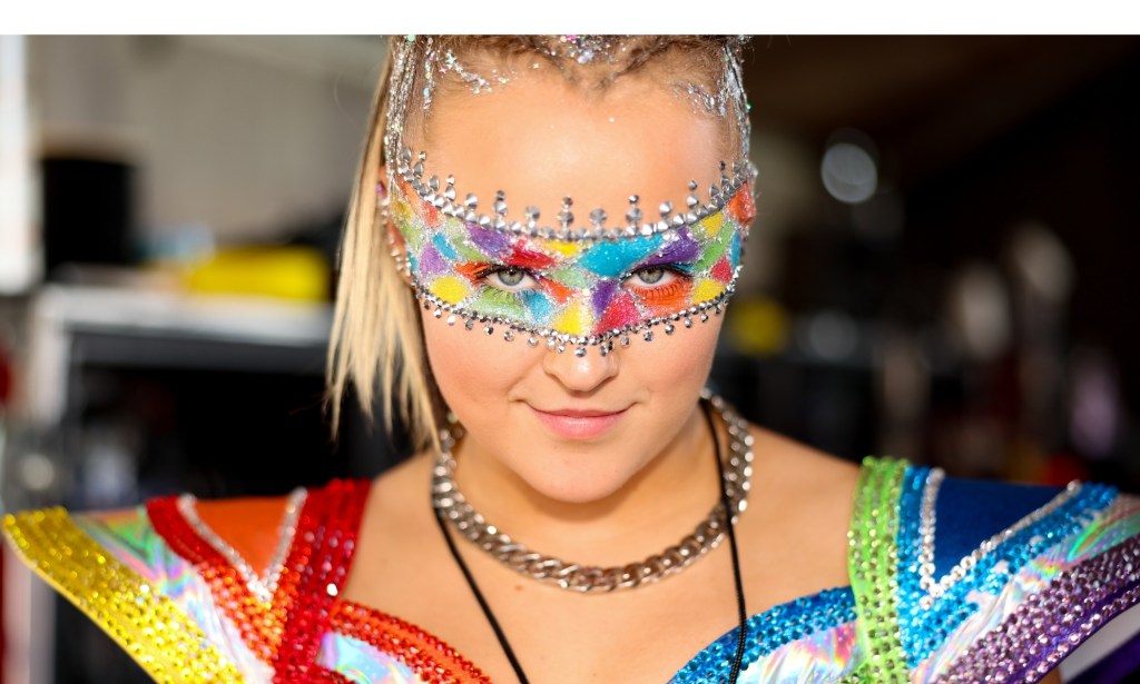 JoJo Siwa dressed in a rainbow concert outfit and rainbow makeup band across her eyes.