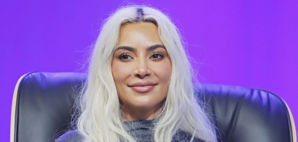 Kim Kardashian on stage, sat on a black chair with a purple background. She's smiling with white hair and a grey high-neck top.