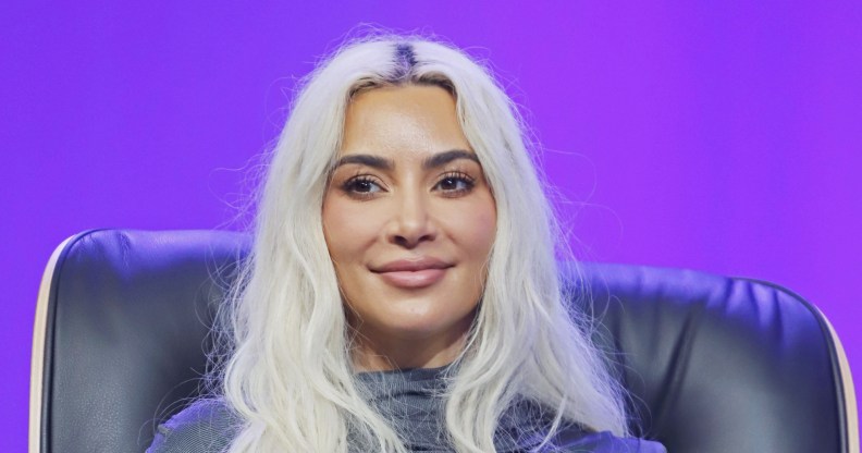 Kim Kardashian on stage, sat on a black chair with a purple background. She's smiling with white hair and a grey high-neck top.