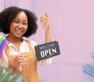 This is an image of a Black woman holding an "we're open" sign in one hand and a Pride flag in the owner. She is wearing a brown apron.