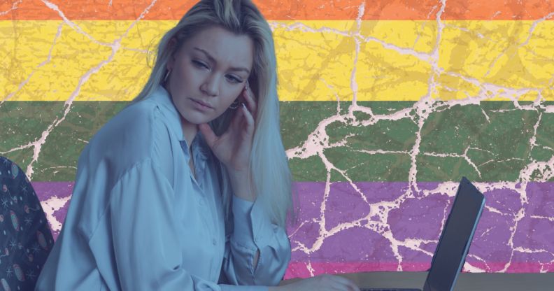 this is a stylized image of a woman looking unhappy or distressed at a computer. She is in a blue tint and behind her is the pride flag, but it is cracked.