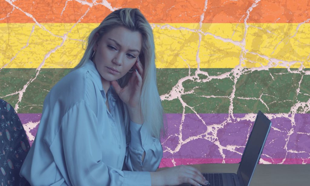 this is a stylized image of a woman looking unhappy or distressed at a computer. She is in a blue tint and behind her is the pride flag, but it is cracked.