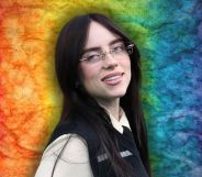 Billie Eilish looks off to the right and smiles. She is wearing glasses and a beige shirt and black vest. She is against a rainbow background.