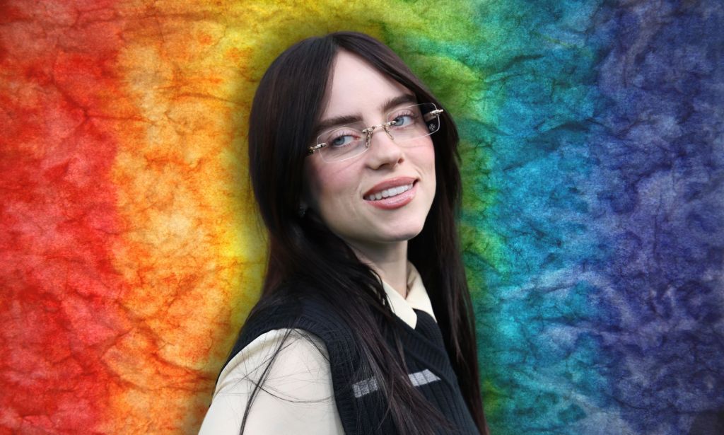 Billie Eilish looks off to the right and smiles. She is wearing glasses and a beige shirt and black vest. She is against a rainbow background.