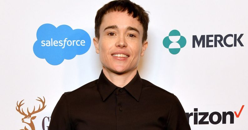 Elliot Page smiles on a red carpet while wearing a dark buttoned shirt.