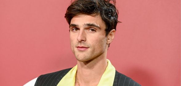 Jacob Elordi standing against a pink background posing. He is wearing a yellow shirt and a pinstriped blazer.