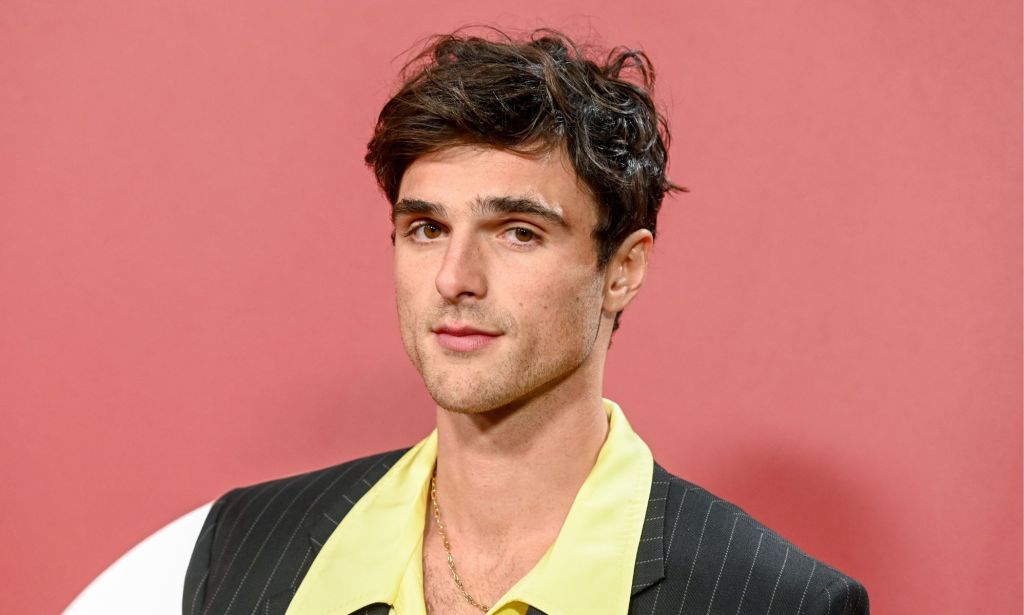 Jacob Elordi standing against a pink background posing. He is wearing a yellow shirt and a pinstriped blazer.