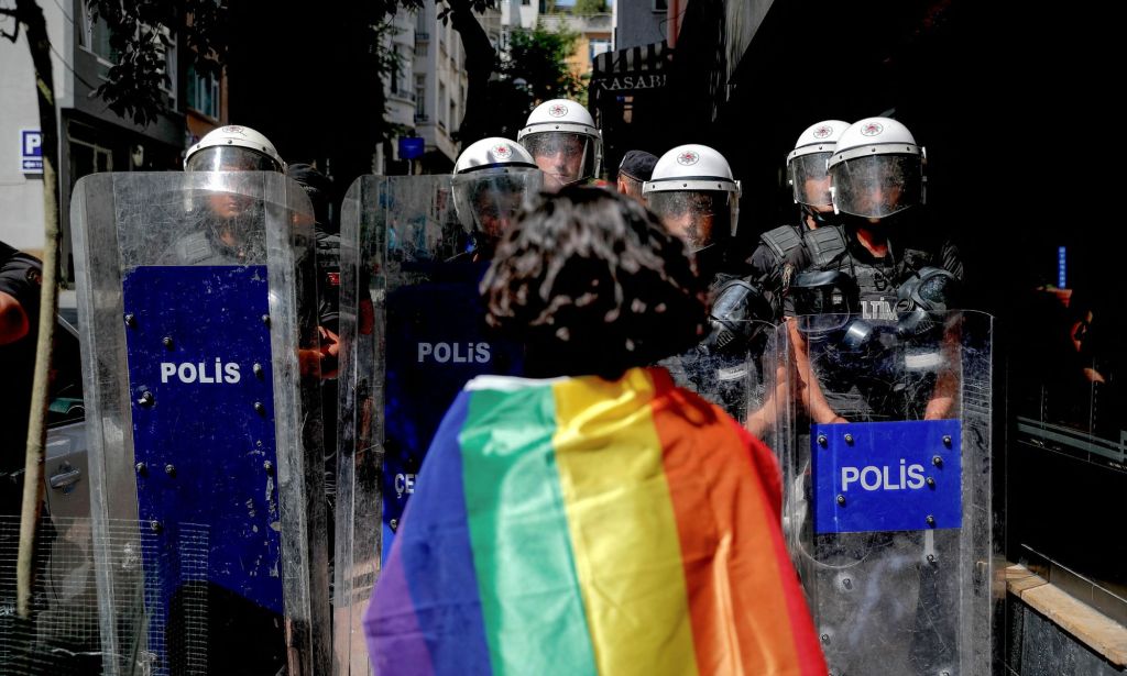 An LGBTQ+ person with a Pride flag on their shoulders standing up to police holding riot shields in Turkey.