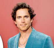 Matt Bomer in a blue blazer and white shirt with silver chain poses for photos against an reddish background.