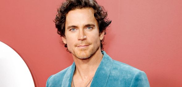 Matt Bomer in a blue blazer and white shirt with silver chain poses for photos against an reddish background.