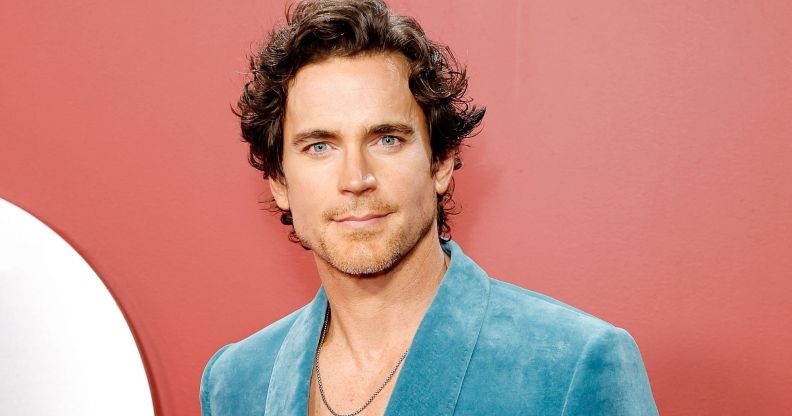 Matt Bomer in a blue blazer and white shirt with silver chain poses for photos against an reddish background.