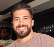 Streamer Nickmercs smiling while wearing a brown top, gold chain and standing in a bar.