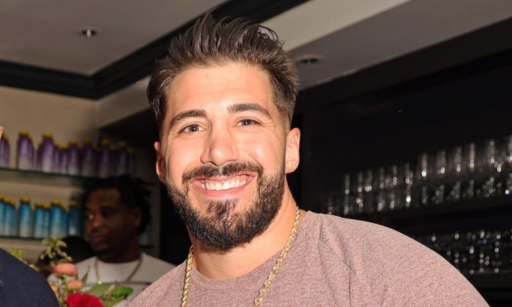 Streamer Nickmercs smiling while wearing a brown top, gold chain and standing in a bar.