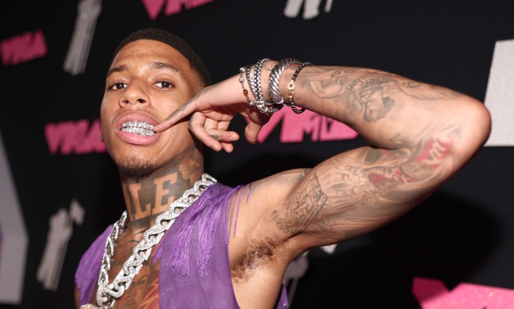 NLE Choppa points to his grill while wearing a chain and purple vest on a red carpet.