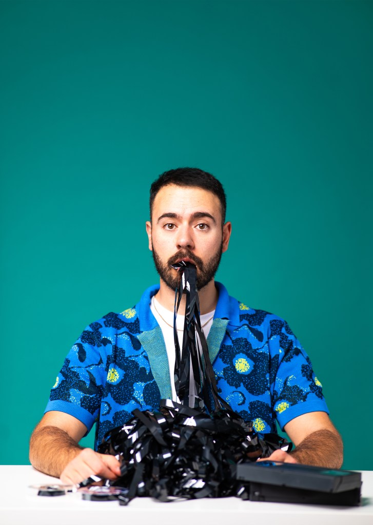 Anesti Danelis: Artificially Intelligent. Anesti is sat at a desk, wearing a blue floral top with VHS tape coming out of his mouth and piling up on the table in front of him.