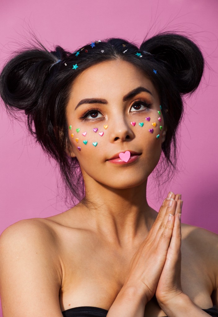 Anna Akana: It Gets Darker. Anna with two spacebuns and colourul hear stickers across her cheeks like freckles and a bigger pink heart on her closed lips.