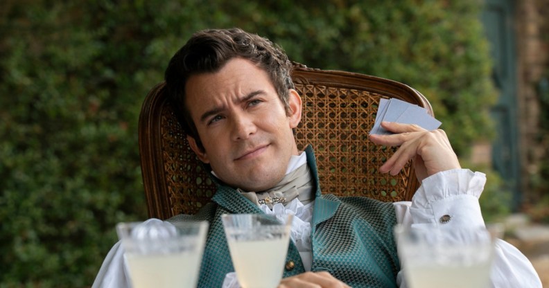 Benedict Bridgerton sitting in a wooden chair with cards and drinks in front of him