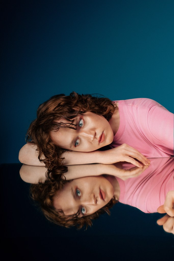 Dee Allum at Edinburgh Fringe. Dee is wearing a pink tshirt lying on a mirrored table. Her mirror reflection is looking at the camera.