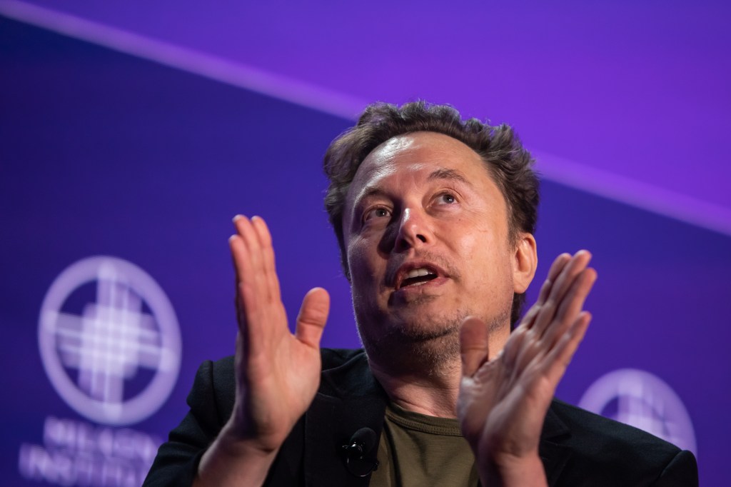 Elon Musk on stage at a panel holding his hand up as he speaks