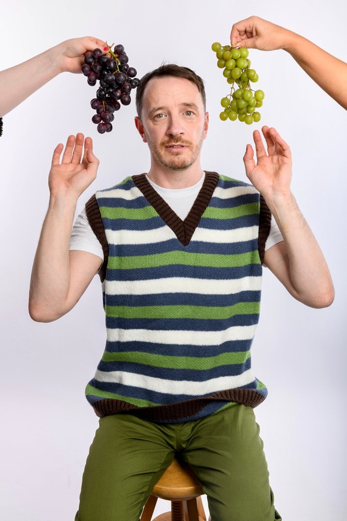 Gearóid Farrelly: Gearóid Rage. Gearóid is sat on a stall with red and whtie grapes about his head. He's wearing a striped white, blue and green sweater vest.