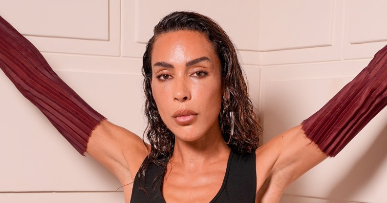 Ines Rau posing against white paneling, she's wearing maroon-brown arm gloves and a black sleevless v-neck top.