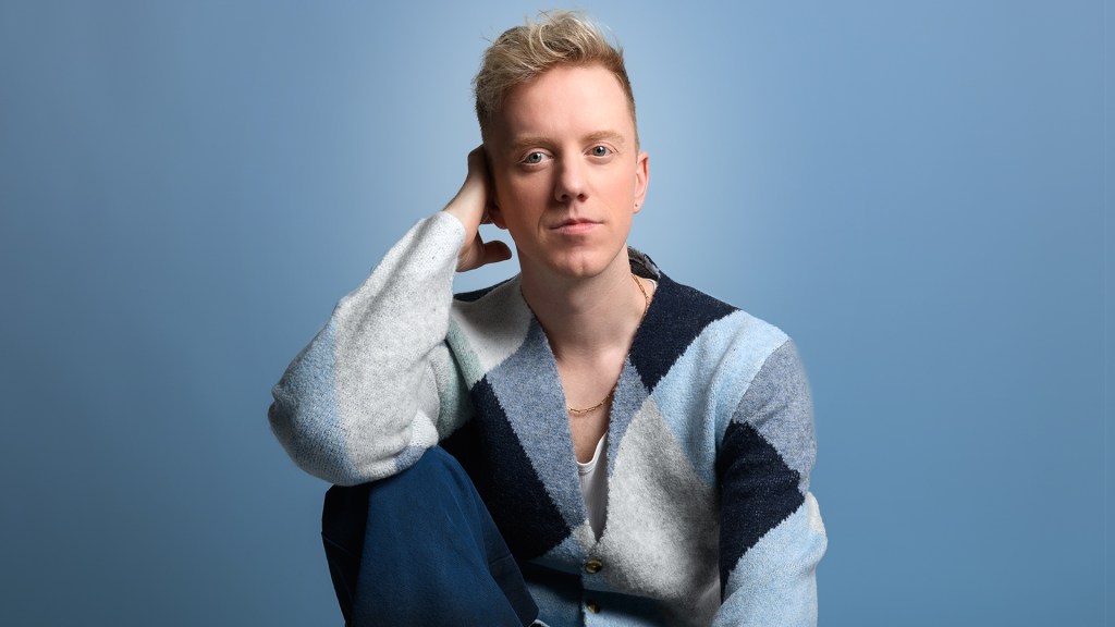 James Barr: Sorry I Hurt Your Son (Said My Ex To My Mum) at Edinburgh Fringe. James is sat against a blue background with a grey, black and blue patchwork jumper.