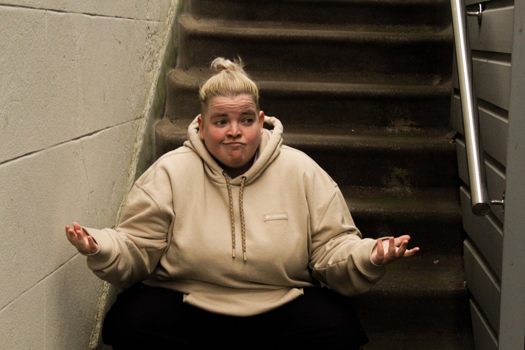 Kim Blythe: Might As Well at Edinburgh Fringe. Kim is wearing a light brown hoodie while sat at the bottom of a stairway.