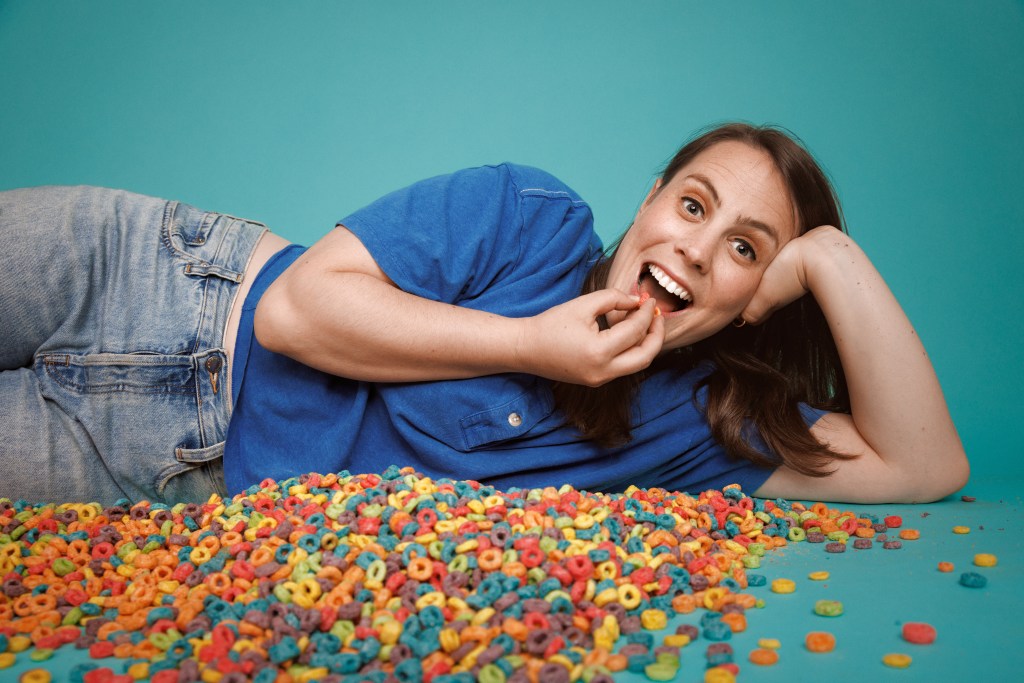 Olivia Levine: Unstuck. Olivia lyng on the ground against a light blue background with cheerios all over the floor, she's eating a handfull.