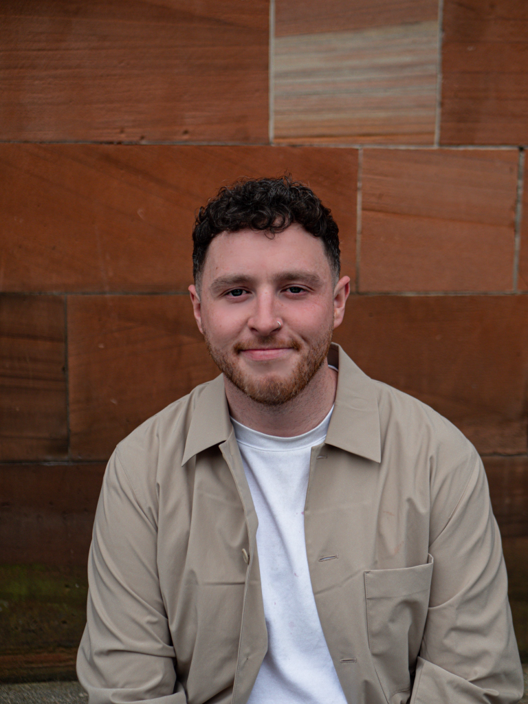 Paul Black: All Sorts at Edinburgh Fringe. Paul is wearing a white tshirt, light brown over jacket while sat against a brick wall