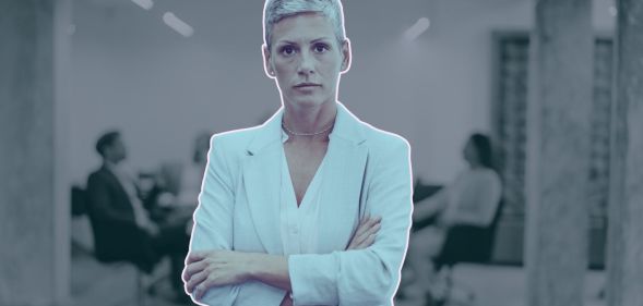 This is an image of an LGBTQ+ professional. She is a woman and she looks frustrated and her arms are folded. In the backgroud there are people in a meeting room.