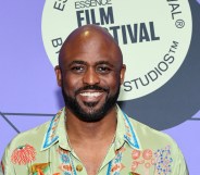 Wayne Brady smiling on a red carpet against a purple background and a floral design green shirt.
