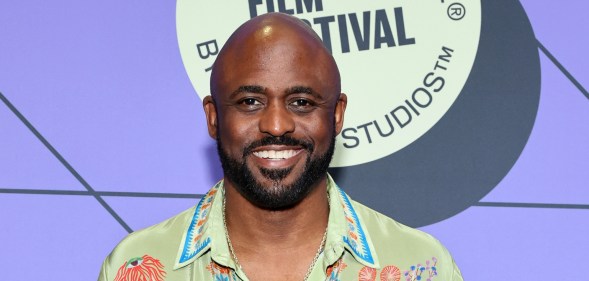 Wayne Brady smiling on a red carpet against a purple background and a floral design green shirt.