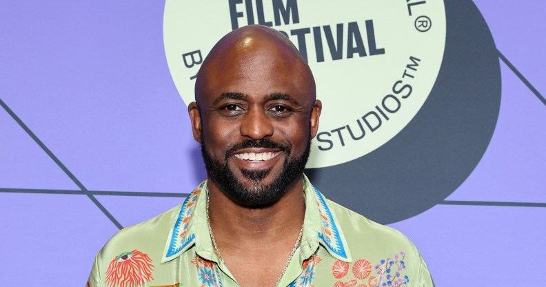 Wayne Brady smiling on a red carpet against a purple background and a floral design green shirt.