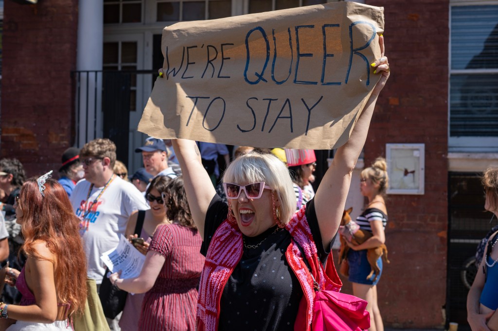 A protester holds a banner reading 'we're queer to stay'.