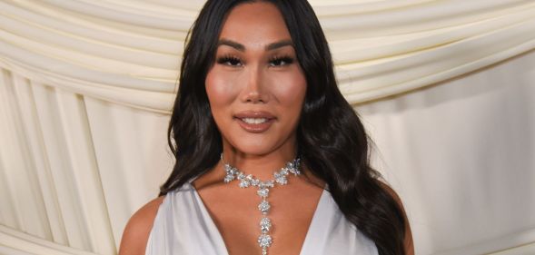 Gia gunn smiles as she wears a white dress and silver necklace and stands against a white curtain