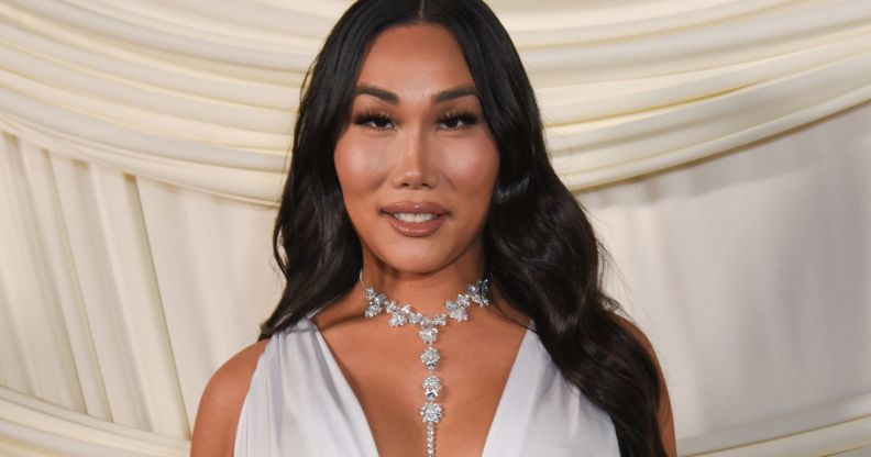 Gia gunn smiles as she wears a white dress and silver necklace and stands against a white curtain