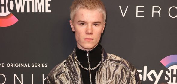 Young Royals star Edvin Ryding in a silver jacket, black jumper and necklace stood against a grey background.