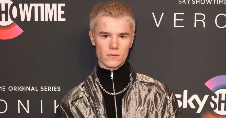 Young Royals star Edvin Ryding in a silver jacket, black jumper and necklace stood against a grey background.