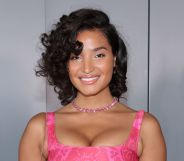 Indya Moore smiles and poses while wearing a pink dress and standing against a grey background.