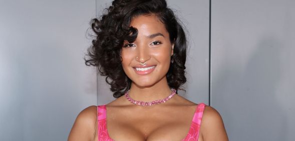 Indya Moore smiles and poses while wearing a pink dress and standing against a grey background.