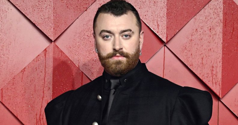 Sam Smith in a black suit posing against a red background.