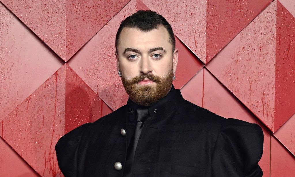 Sam Smith in a black suit posing against a red background.