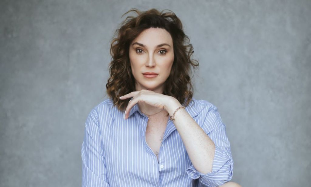 Juno Dawson poses with her hand on her chin, wearing a blue shirt, against a grey background.