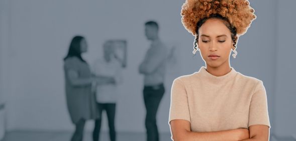 This is an image of a Black woman looking stressed and isolated at work. She has her arms crossed. In the background there are 3 other people blurred.