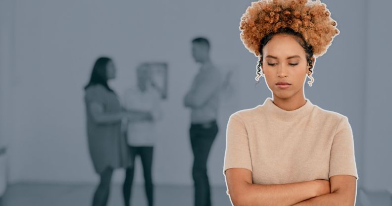 This is an image of a black woman looking stressed and isolated at work. She has her arms crossed. Three other people are visible in the background, out of focus.