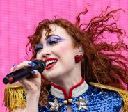 Chappell Roan performing against a bright pink background. She is wearing a blue sequin and gold fringe shirt with big red earrings