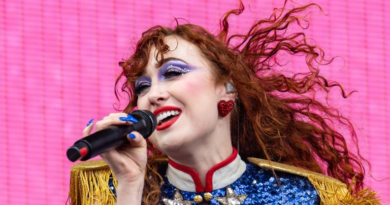 Chappell Roan performing against a bright pink background. She is wearing a blue sequin and gold fringe shirt with big red earrings