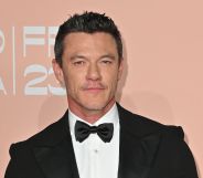 Luke Evans in a black tuxedo and bow tie against a peach background